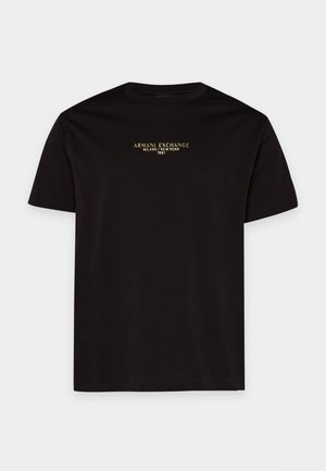 Un homme porte un tee-shirt noir à logo, un jean foncé, des baskets noires et une casquette blanche sur un fond clair et uni.