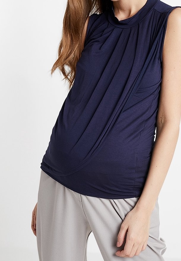 Femme portant un top sans manches bleu marine drapé et un pantalon gris clair, debout devant un fond blanc uni avec une main reposant sur la cuisse.