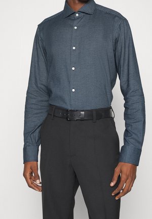 Man draagt een donkerblauw patroon overhemd met lange mouwen, ingestopt in een zwarte broek met een zwarte leren riem.