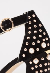 Chaussure à bride cheville en daim noir avec clous dorés et ornements en perles, munie d'une boucle ronde en or.