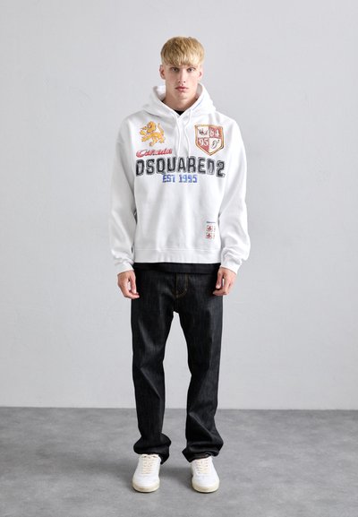 Witte hoodie met grafische afdrukken en patches, gecombineerd met donkere jeans en witte sneakers. De hoodie heeft een leeuw en logo-details.