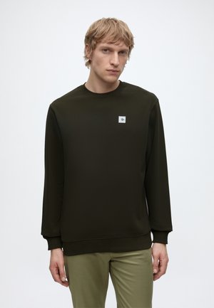 Junger Mann mit kurzen blonden Haaren, der einen dunkelgrünen Sweatshirt mit langen Ärmeln und hellgrüne Hose trägt, steht vor einem schlichten weißen Hintergrund.