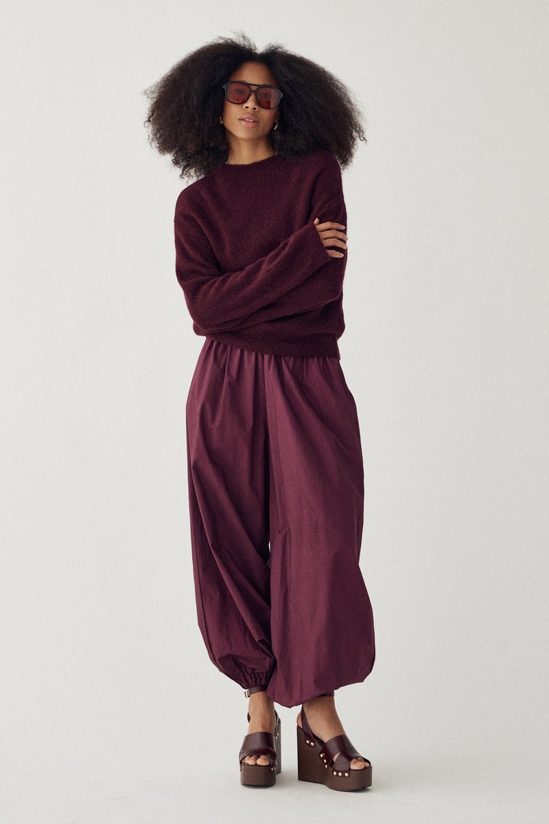 Žena s veľkými kučeravými vlasmi, oblečená v tmavočervenom svetri, voľných bordových nohaviciach, platformových sandáloch a slnečných okuliaroch, stojaca so založenými rukami.