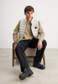 Helle beige gesteppte Weste mit schwarzen Akzenten und einem Logo-Patch; kombiniert mit einem beigen Sweatshirt und dunklen Hosen. Schwarze klobige Stiefel.