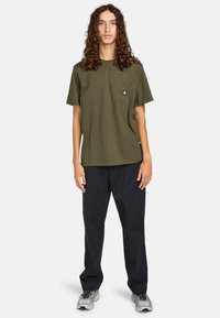Olive green cotton t-shirt with a chest pocket, short sleeves, and a relaxed fit, paired with black trousers and gray sneakers.