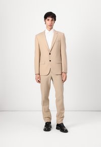 Costume beige à un bouton, avec deux boutons noirs et des revers en pointe. Associé à une chemise blanche et des chaussures noires, sur un fond uni.