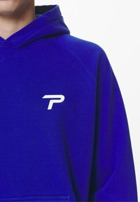 Modrá mikina s kapucí vyrobená z materiálu jako je sweatshirt, s bílým logem "P" na hrudi, se šňůrkami na kapuci a žebrovaným lemem.
