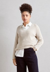 Beżowy sweter dzianinowy z dekoltem w serek, noszony na białej koszuli z kołnierzykiem; zestawiony z ciemnymi spodniami. Posiada długie rękawy i luźny krój.