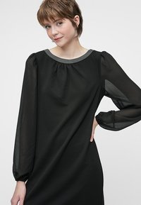 Black dress made of lightweight fabric, featuring a round neckline with a beaded trim and puffed long sleeves. Smooth texture and flowing design.