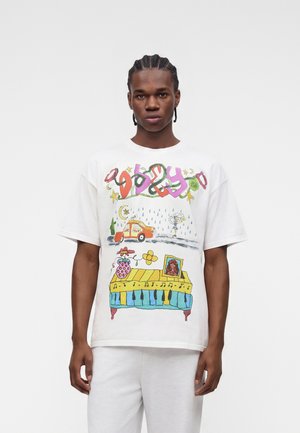 Jeune homme aux cheveux tressés portant un t-shirt blanc avec des dessins colorés d'une voiture, d'un piano, d'une fleur et de formes abstraites.
