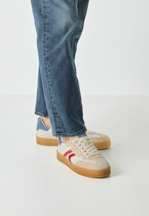 Zapatillas deportivas que presentan una parte superior beige de ante y textil, detalles blancos, franjas rojas y un talón azul, con una suela de goma color marrón claro.
