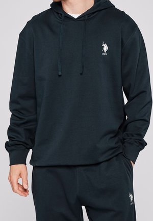 Mężczyzna ubrany w czarną bluzę z kapturem USPA i dopasowane spodnie dresowe, prezentujący biały logotyp polo gracza na klatce piersiowej i udzie, stoi z jedną ręką w kieszeni.