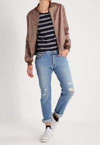 Chaqueta bomber marrón sobre una camisa de rayas en azul y blanco, combinada con jeans claros desgastados y zapatillas blancas. Atuendo casual.