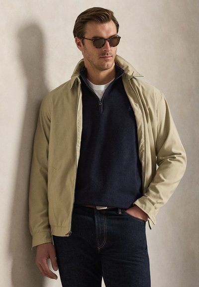 Homme portant une veste beige par-dessus un pull marine avec fermeture éclair quart, jean foncé et lunettes de soleil, debout contre un mur beige uni.