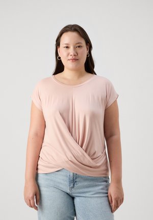 Light pink short-sleeve shirt with a twisted front design. Made of soft fabric, featuring a round neckline and relaxed fit. Worn with blue jeans.