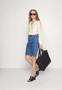 Blusa de manga longa em tom de creme claro, calções bermuda em denim azul, mules pretos de dedos abertos e uma mala de mão texturizada preta com alças duplas.