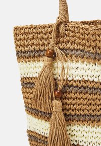 Sac fourre-tout en paille avec une texture tressée, présentant des rayures horizontales en brun, crème et beige. Équipé de pompons décoratifs et de perles en bois.