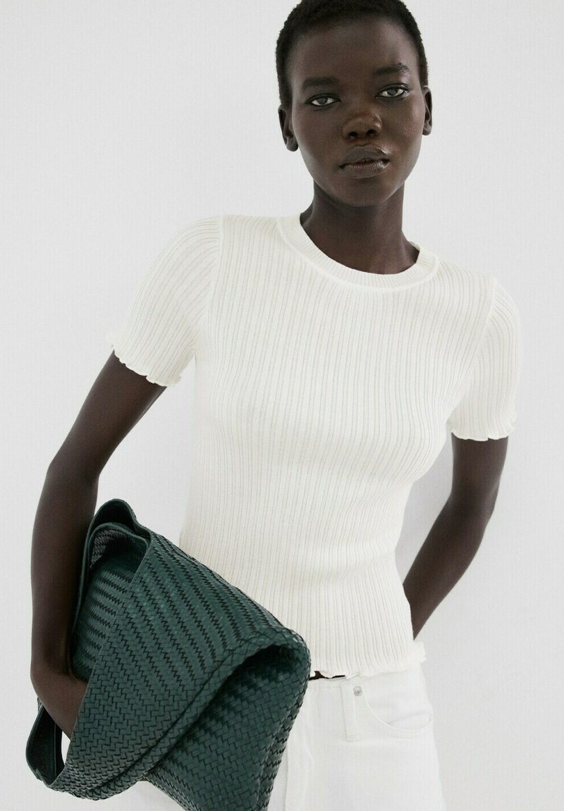 Kobieta ubrana w biały prążkowany top z krótkim rękawem i białe spodnie, trzymająca dużą ciemnozieloną plecioną torbę na jednolitym tle.