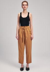 Zwarte geribbelde tanktop gecombineerd met hoog-zittende, mosterdkleurige broeken met een strik, knoopzakken en een rechtlopende pasvorm.