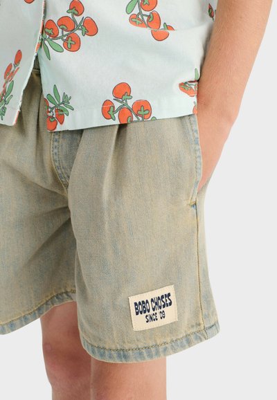 Child wearing light blue denim shorts with "BOBO CHOSES SINCE '09" patch, hand in pocket, and a light shirt with red tomato prints.