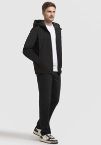 Sort letvægtsjakke med hætte, lynlås foran og sidelommer; båret over en hvid t-shirt og sorte jeans, parret med sneakers.