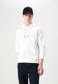 Hvid hættetrøje med snore og frontlomme, parret med mørke jeans. Modellen bærer en sort kasket og står foran en lys baggrund.