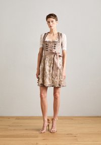 Robe dirndl en beige avec un haut en dentelle, un corsage cintré, des broderies florales sur la jupe et un nœud en satin. Sandales à talons hauts transparentes.