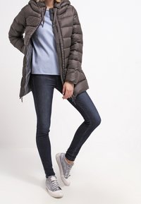 Donkergrijze gewatteerde jas met een capuchon, ritsclosure en doorstikte design, gedragen over een lichtblauw shirt en donkere jeans, in combinatie met grijze sneakers.