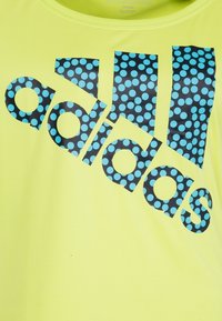 Neongul t-shirt med svart "adidas"-logotyp fylld med blå prickar, korta ärmar och rund halsringning.