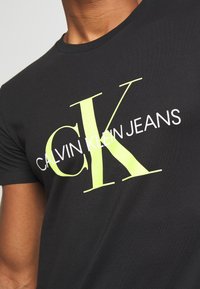 Czarny t-shirt z dużymi neonowo-żółtymi literami "CK" oraz białym napisem "Calvin Klein Jeans" na piersi.