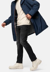 Manteau matelassé bleu marine avec accents noirs, chemise beige clair, jean noir et bottines crème lacées avec semelles épaisses, présenté en position de marche.