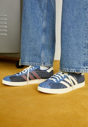 Osoba nosząca niebieskie buty sportowe Adidas Gazelle z wzorzystym wzorem i białymi sznurówkami, zestawione z niebieskimi dżinsami na musztardowym dywanie.