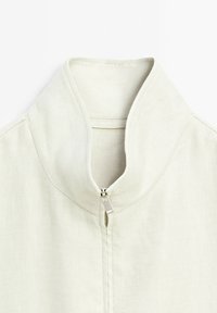 Giacca bianca con zip e colletto alto. Presenta una texture liscia e un sottile motivo a trama diagonale. Dettaglio della zip argentata sul davanti.