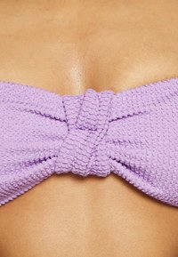 Top de bikini roxo texturizado com detalhe central em nó, apresentando um padrão de borda ondulada e uma superfície de tecido suave.