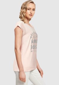 Lys rosa t-skjorte med korte oppbrettede ermer, V-hals og et trykt grått tekstdesign på forsiden. Kombinert med hvite bukser.