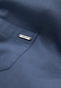 Tessuto strutturato blu navy con un piccolo tag metallico argentato etichettato "OMBRIE DEPT." fissato con due ribattini vicino a una cucitura della tasca.