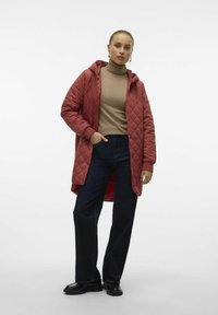 Manteau matelassé à capuche couleur rouge rouille, associé à un col roulé beige et à un jean large bleu foncé. Texture lisse et coupe à mi-mollet.