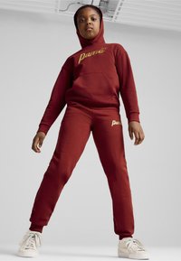 Conjunto de sudadera y pantalones burdeos con logo dorado, que presenta una capucha, bolsillo frontal y puños elásticos, estilizado en un ajuste relajado.