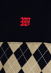 Haut en tissu noir avec un motif brodé rouge, se transformant en une section beige à motifs présentant des losanges noirs alternés et des coutures blanches.