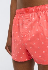 Κόκκινο βαμβακερό μπόξερ με ελαστική ζώνη μέσης, διακοσμημένο με επαναλαμβανόμενα λευκά λογότυπα Calvin Klein σε όλο το ύφασμα.