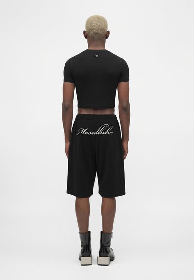 Άτομο με κοντά ξανθά μαλλιά στέκεται με την πλάτη, φορώντας μαύρο crop top, μαύρα σορτς με το κείμενο «Masallah» και μαύρες μπότες με λευκές σόλες.