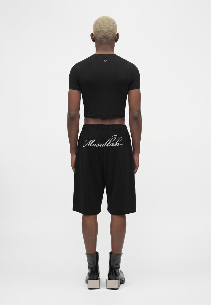 Person with short blond hair stands facing away, wearing black crop top, black shorts with "Masallah" text, and black boots with white soles.