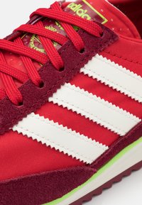 Röd tygsneaker med en texturerad bourgogne mockaöverlappning, med tre vita ränder, röda snören och en grön accent på sulan.