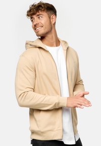 Beige zip hoodie med ribkant i bunden og ved håndleddene, med en hætte med snoretræk og logo accent på halsudskæringen. Båret over en hvid skjorte.