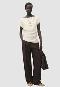 Haut blanc texturé avec un large col, associé à un pantalon ample marron foncé. Comprend un collier pendentif en or et un sac fourre-tout marron.