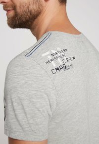 Szary T-shirt z krótkim rękawem, z akcentami w postaci przeszyć, okrągłym dekoltem i nadrukowanym tekstem z tyłu. Tkanina ma lekką teksturę.
