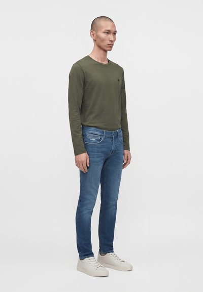Homme debout portant une chemise olive à manches longues, un jean slim bleu et des baskets blanches sur un fond clair uni.