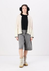 Cremefarbener Cardigan mit schwarzem Saum, schwarzes T-Shirt, graue Denim-Shorts und beigefarbene, gefütterte Stiefel. Das Modell steht vor einem schlichten weißen Hintergrund.