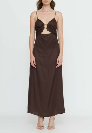 Donna che indossa un vestito lungo marrone scuro senza maniche con sottili bretelle, un dettaglio circolare tagliato sul davanti e tacchi aperti dorati.