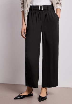 Frau trägt schwarze, weit geschnittene Hosen mit Gürtel, beige Blazer, weißes Top und schwarze Slingback-Ballerinas und steht drinnen.
